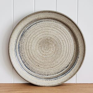 May include: A round, speckled ceramic plate in a light beige hue, with a dark blue accent ring. The plate has concentric circular grooves, giving it a textured surface. It's set against a white and wood backdrop.
