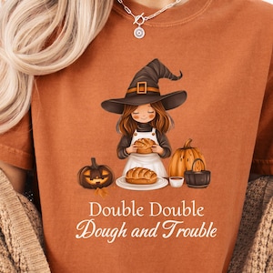 May include: A rust-colored t-shirt featuring a graphic of a witch baking bread with pumpkins and a cauldron. The text "Double Double Dough and Trouble" is printed below the illustration in a white font. The shirt has a crew neck and short sleeves.