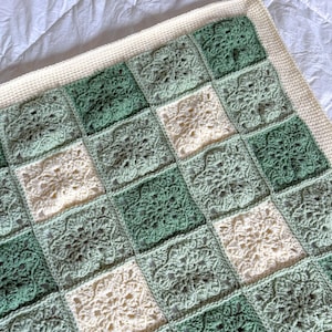May include: A handcrafted crochet blanket with a patchwork design of green and cream squares. Each square features a floral pattern, and the blanket has a cream border. The blanket is soft and cosy.