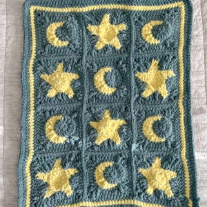 May include: A crocheted baby blanket with a blue background and yellow stars and moons. The blanket has a yellow border.