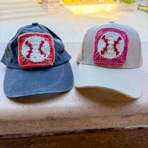 May include: Two baseball caps, one dark grey and one beige, each featuring a square crochet patch on the front. The patches depict a white baseball with red stitching, framed by a border in either muted red or bright pink yarn.