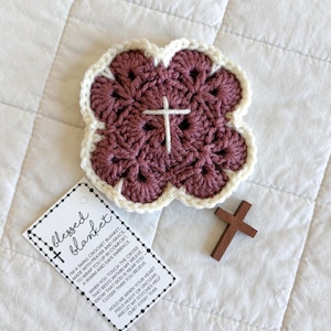 May include: A crocheted blanket in a four-leaf clover shape, with a white cross in the center. The blanket is a deep mauve color with a cream-colored border. A small wooden cross and a tag that says "blessed blanket" are also in the image.