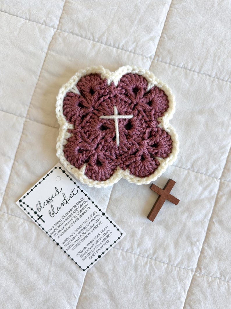 May include: A crocheted blanket in a four-leaf clover shape, with a white cross in the center. The blanket is a deep mauve color with a cream-colored border. A small wooden cross and a tag that says "blessed blanket" are also in the image.