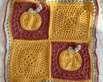 May include: A handmade crochet square blanket featuring four squares in shades of yellow and burgundy. Two squares have a pumpkin applique. The blanket has a light green and peach border. The blanket is on a white surface.