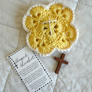 May include: A yellow crochet blanket in a flower shape with a white cross stitched in the center. A small wooden cross and a tag that reads "blessed blanket" are also in the image.
