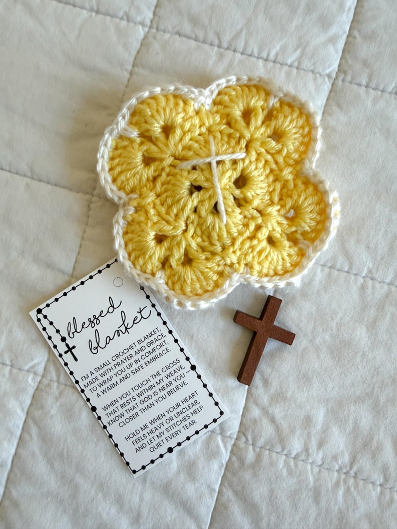 May include: A yellow crochet blanket in a flower shape with a white cross stitched in the center. A small wooden cross and a tag that reads "blessed blanket" are also in the image.