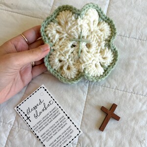 May include: A cream-colored, crocheted flower-shaped blanket with a light green border. A small wooden cross and a tag that reads "blessed blanket" are also in the image. The blanket is designed to offer comfort and prayer.