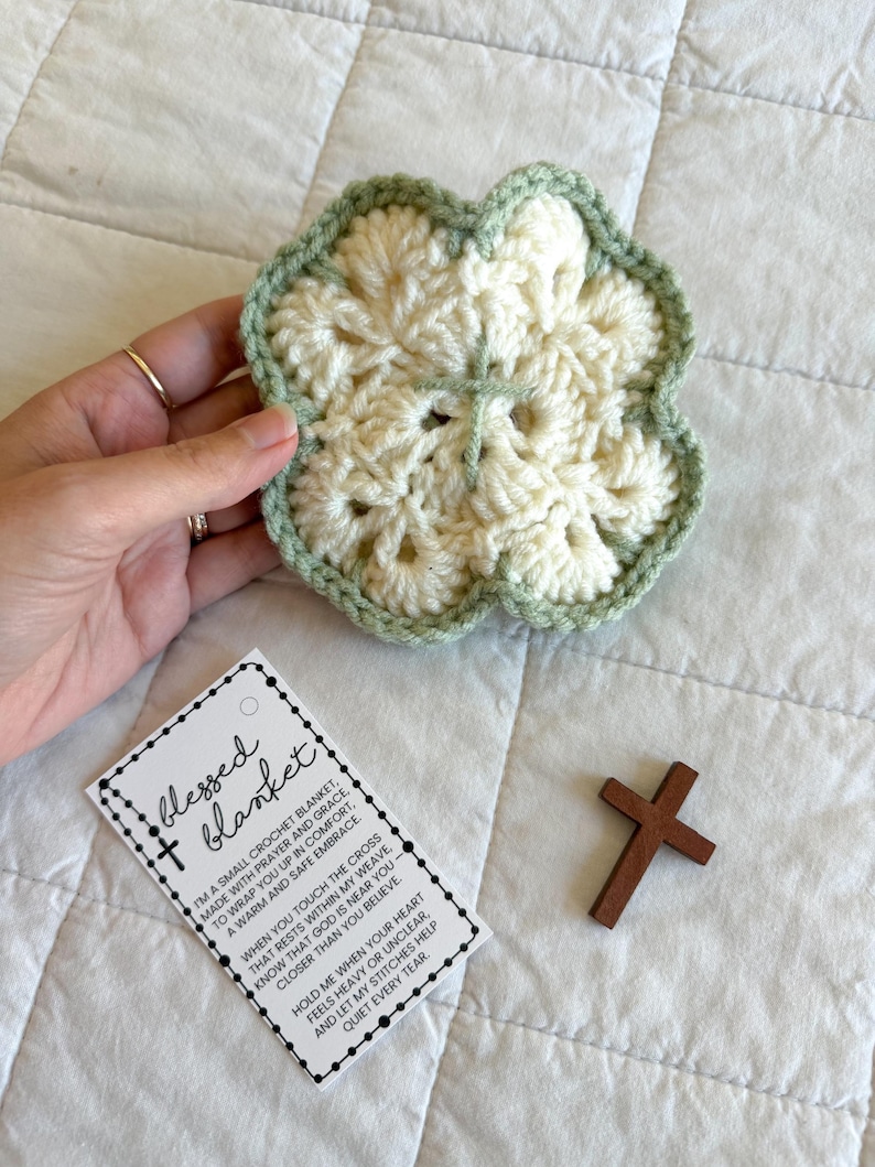 May include: A cream-colored, crocheted flower-shaped blanket with a light green border. A small wooden cross and a tag that reads "blessed blanket" are also in the image. The blanket is designed to offer comfort and prayer.