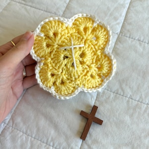 May include: A handmade, yellow crochet coaster in a flower shape with a white border and a white cross stitched in the center. A small, brown wooden cross rests nearby. The coaster is held by a hand.