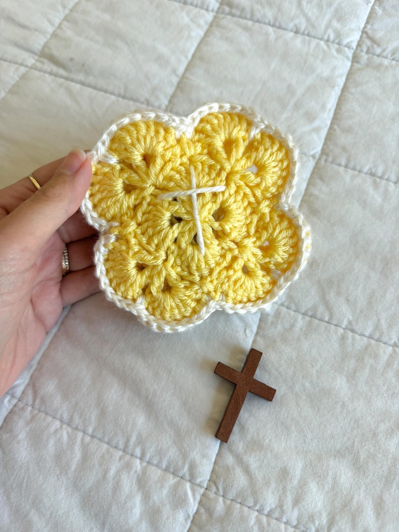May include: A handmade, yellow crochet coaster in a flower shape with a white border and a white cross stitched in the center. A small, brown wooden cross rests nearby. The coaster is held by a hand.