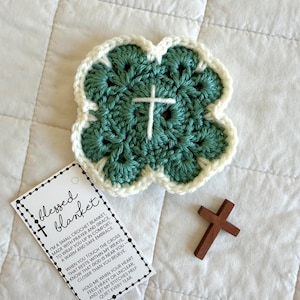 May include: A handmade crochet blanket in a four-leaf clover shape, with a white cross in the center. The blanket is teal with a white border. A small wooden cross and a tag that reads "blessed blanket" are also present.