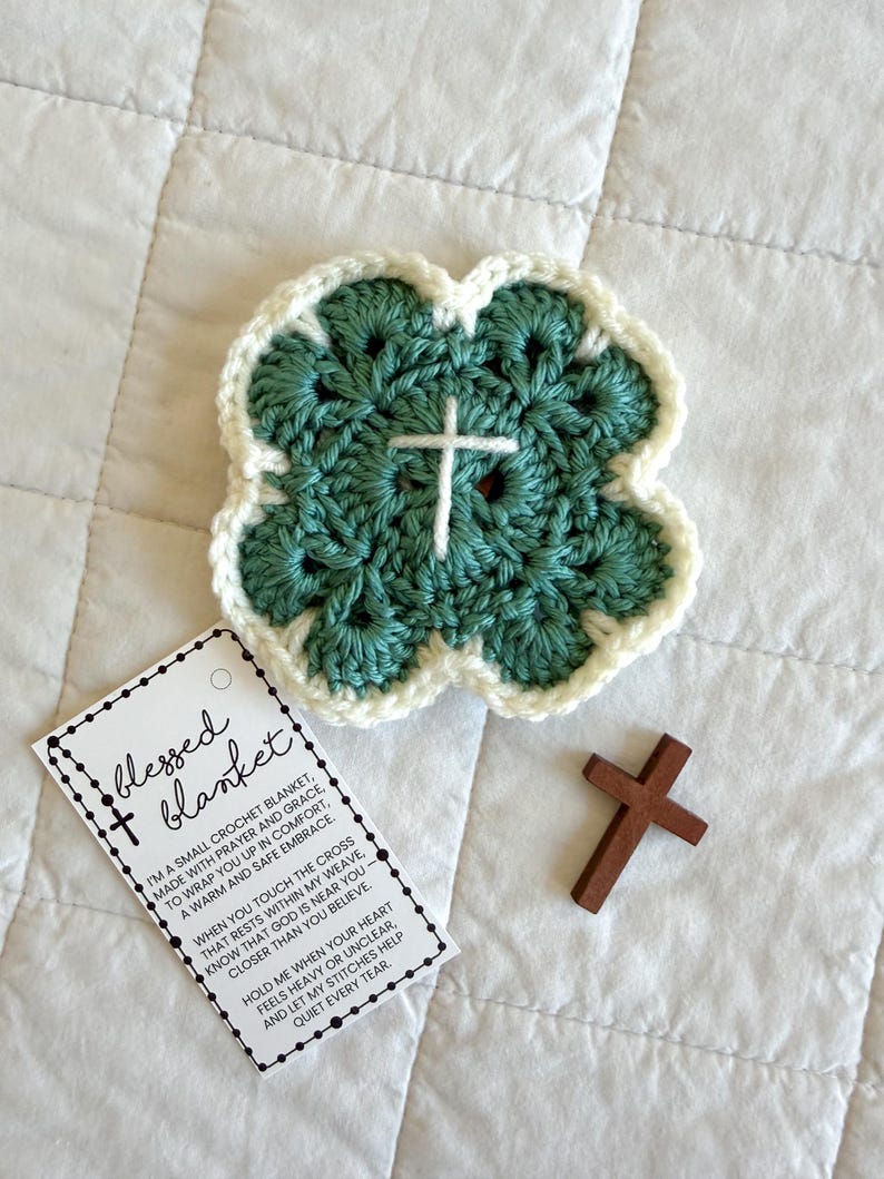 May include: A handmade crochet blanket in a four-leaf clover shape, with a white cross in the center. The blanket is teal with a white border. A small wooden cross and a tag that reads "blessed blanket" are also present.