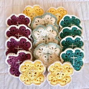 May include: A collection of handmade crochet coasters in various colors, including yellow, purple, green, and cream. Each coaster features a four-petal flower design with a white cross in the center and a white scalloped edge. The coasters are arranged on a white quilted surface.
