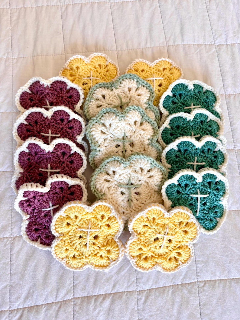 May include: A collection of handmade crochet coasters in various colors, including yellow, purple, green, and cream. Each coaster features a four-petal flower design with a white cross in the center and a white scalloped edge. The coasters are arranged on a white quilted surface.