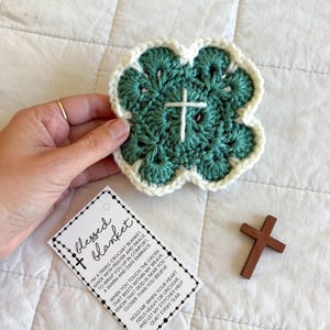 May include: A handmade, four-leaf clover-shaped crochet blanket in teal and cream, with a white cross in the center. A small wooden cross and a tag that reads "blessed blanket" are also present.