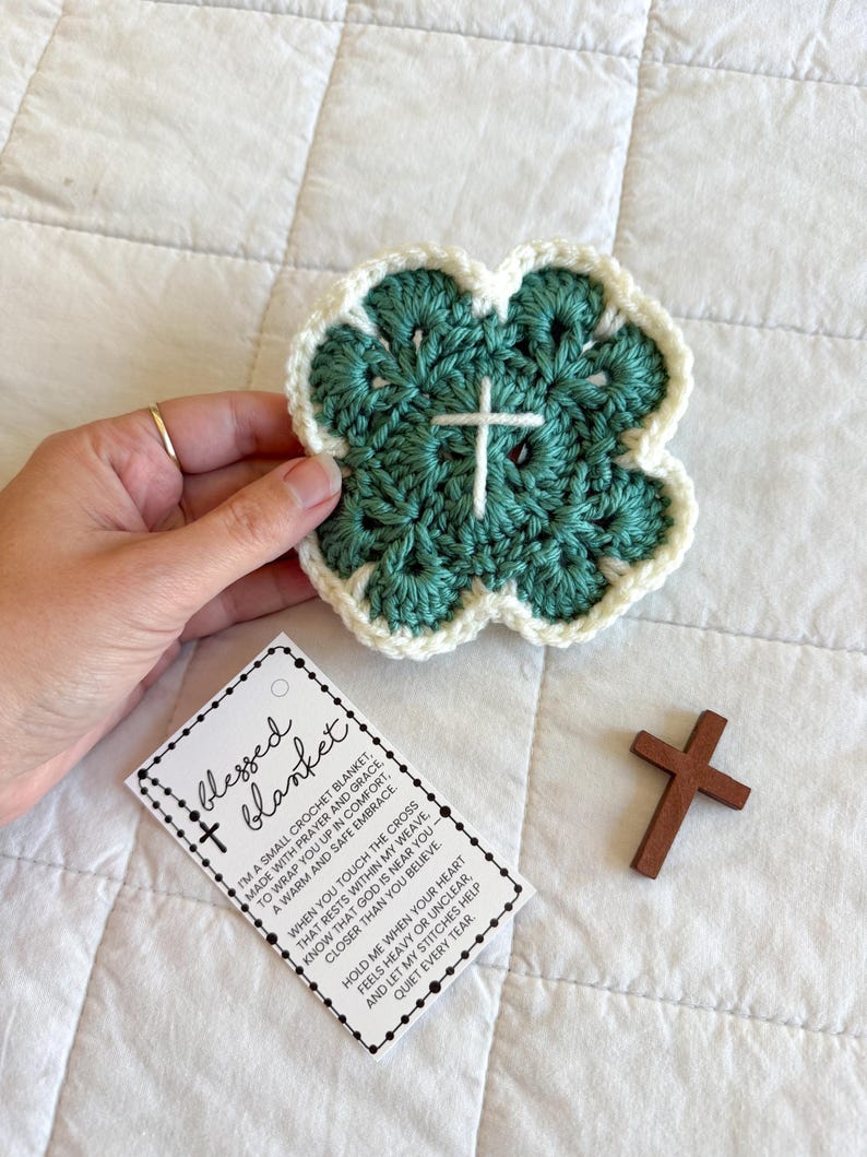 May include: A handmade, four-leaf clover-shaped crochet blanket in teal and cream, with a white cross in the center. A small wooden cross and a tag that reads "blessed blanket" are also present.