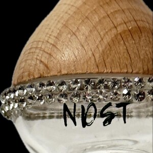 Könnte beinhalten: Eine klare Glasflasche mit einem Holzdeckel und einem Band aus funkelnden Strasssteinen. Das Wort "NOST" ist in schwarzer Schrift auf dem Glas geschrieben. Die Flasche steht vor einem schwarzen Hintergrund.