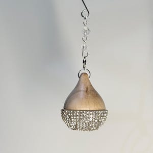 Könnte beinhalten: Tropfenförmiger Anhänger mit Holzoberteil und einer mit glitzernden Strasssteinen besetzten Unterseite. Der Anhänger hängt an einem silbernen Draht mit klaren Kristallperlen. Der Hintergrund ist ein weiches Grau.