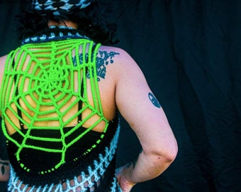 Könnte beinhalten: Eine grüne, gehäkelte Spinnennetz-Weste mit einem schwarz-weißen Häkelmuster. Die Weste wird über einem schwarzen Tanktop getragen.