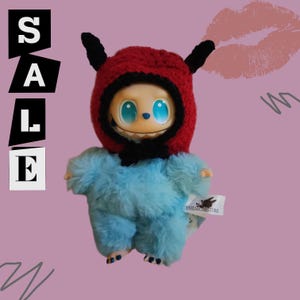 May include: A plush toy with a blue body and a red crocheted hood with black horns. The toy has large blue eyes and a smiling face. The word "SALE" is displayed in white block letters on the left side of the image.