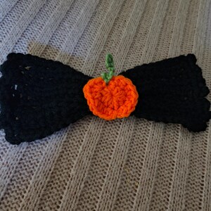 Op de afbeelding: Een zwarte gehaakte strik met een oranje gehaakte pompoen in het midden. De pompoen heeft een groene stengel. De strik ligt op een lichtgrijze gebreide ondergrond. Dit handgemaakte item is perfect voor Halloween.