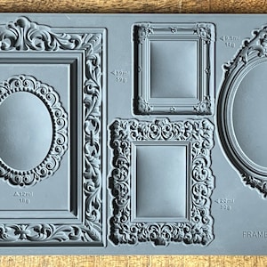 May include: A gray silicone mold featuring five ornate picture frame designs. The frames vary in shape and size, including rectangular and oval options. The mold is labeled "FRAMES IOD" and includes volume and weight measurements.