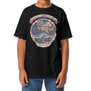 Peut inclure: T-shirt noir avec un motif d'un dinosaure portant des lunettes de soleil et un drapeau am&eacute;ricain, avec le texte "FREEDOMOSAURUS REX" et "EST. 1776". Le dinosaure est sur un monster truck avec un motif de drapeau am&eacute;ricain.