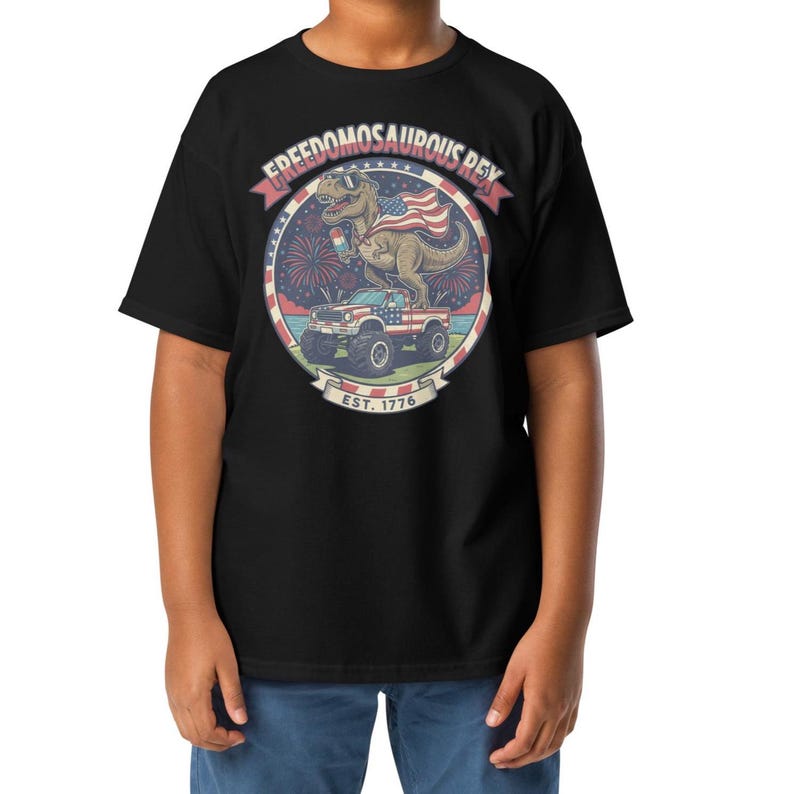 Peut inclure: T-shirt noir avec un motif d'un dinosaure portant des lunettes de soleil et un drapeau am&eacute;ricain, avec le texte "FREEDOMOSAURUS REX" et "EST. 1776". Le dinosaure est sur un monster truck avec un motif de drapeau am&eacute;ricain.