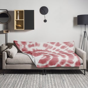 May include: A red and white tie-dye throw blanket is draped over a grey sofa. The blanket has a circular pattern with a white centre and red swirls.
