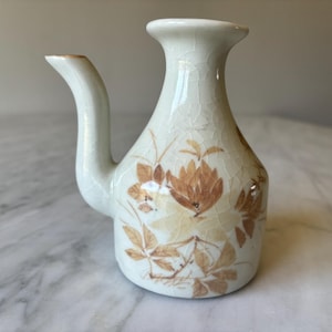 May include: A cream-colored ceramic pitcher with a curved spout and floral design. The pitcher features brown and tan floral patterns and a crackled glaze. The pitcher is approximately 6 inches tall.