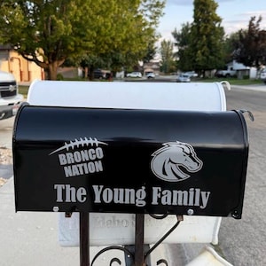 Puede incluir: Buzón negro con el texto "Bronco Nation" y "The Young Family" en blanco. Un logotipo de cabeza de caballo plateado está en el lado derecho. El buzón está montado en un poste de metal con otros dos buzones.