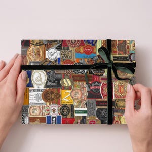 May include: A rectangular gift box wrapped in paper featuring a collage of various colorful cigar bands. The box is tied with a dark green velvet ribbon that forms a bow on the top surface, contrasting with the intricate, multi-colored labels.