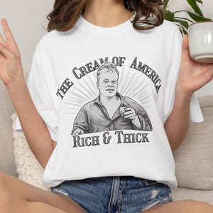 May include: White t-shirt with a graphic that reads "The Cream of America" above an illustration of a man holding a cup, with "Rich & Thick" below. The person is wearing denim shorts.
