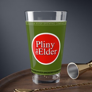 Può includere: Un bicchiere trasparente con liquido verde, con un logo circolare rosso e bianco che recita "Pliny the Elder". Il bicchiere ha una fascia verde con la scritta "Russian River Brewing Company". Un cucchiaio da bar e un jigger dorati sono su un vassoio di legno.