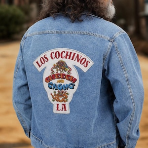 Può includere: Una giacca di jeans azzurra con una grande toppa ricamata sul retro. La toppa mostra il testo LOS COCHINOS, CHEECH AND CHONG e LA, accompagnato da colorate illustrazioni grafiche di figure stilizzate e creature simili a draghi.