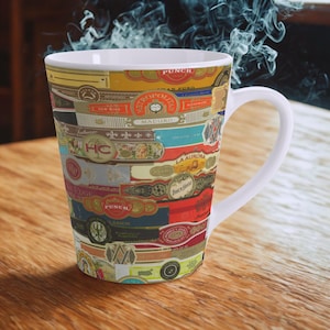 Op de afbeelding: Een witte keramische koffiemok met een collage van kleurrijke sigarenbandjes. De bandjes tonen verschillende merknamen en ontwerpen in rood, goud, blauw en andere kleuren. De mok heeft een gebogen oor en een taps toelopende vorm.