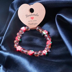 May include: A beaded bracelet featuring translucent pink spherical beads interspersed with small, colorful fruit-shaped charms. The bracelet is attached to a heart-shaped cardboard tag printed with the text Handmade with love Thank you.
