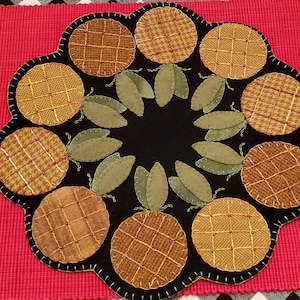 Puede incluir: Un mantel individual artesanal con borde negro festoneado. El diseño presenta hojas verdes y círculos en tonos marrones y amarillos, con patrones texturizados. El mantel individual está sobre una superficie roja. Diámetro aproximado de 30 cm.