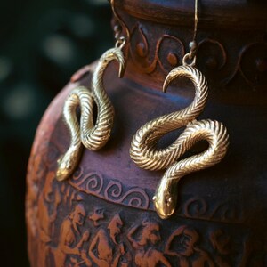 Puede incluir: Un par de pendientes dorados con forma de serpiente. Los pendientes tienen la forma de una serpiente enrollada con escamas detalladas y una cabeza curvada. Cuelgan de un gancho simple. El fondo es un jarr&oacute;n marr&oacute;n borroso.