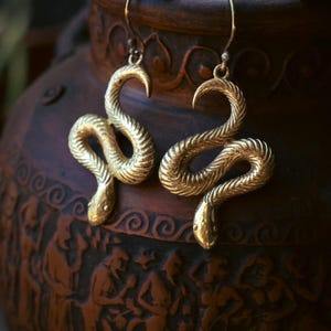 Może przedstawiać: Para złotych kolczyków w kształcie węża. Kolczyki są szczegółowe z łuskami i zakrzywioną głową, zawieszone na prostym haczyku. Tło przedstawia ciemnobrązowy, teksturowany wazon z rzeźbionymi postaciami.