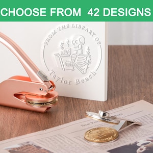Op de afbeelding: Een roségouden reliëfstempel met een cirkelvormige stempel en lange handgreep, naast een witte kaart met een bibliotheekzegelontwerp. De tekst "CHOOSE FROM 42 DESIGNS" staat bovenaan. Een krant en een gouden munt zijn ook in de afbeelding.