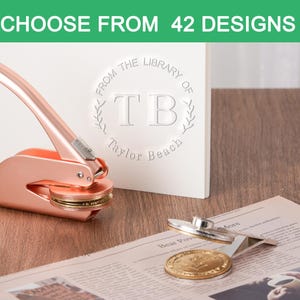 Könnte beinhalten: Ein roségoldener Prägestempel neben einer weißen Karte mit dem Text "FROM THE LIBRARY OF TB Taylor Beach". Der Prägestempel befindet sich auf einer Holzoberfläche, mit einer Zeitung und einem goldenen Prägesiegel.