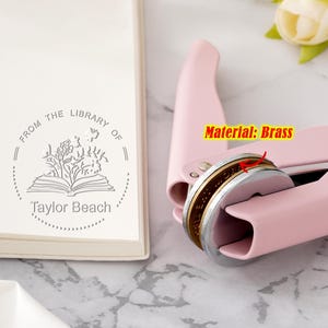 May include: A pink self-inking stamp with a brass stamp head. The stamp reads "From the Library of Taylor Beach" with a stylised image of a book and flowers.