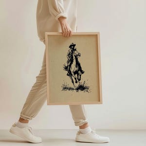 May include: A rectangular wooden frame containing a print on tan paper. The print features a black ink illustration of a person wearing a cowboy hat riding a horse through tall grass. The artwork is centered within the light-colored wooden frame.