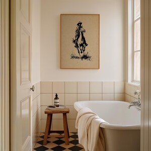 May include: A framed wall art print featuring a black ink illustration of a cowboy riding a horse on a tan background. The artwork is displayed on a wall above a white bathtub and a small wooden stool in a tiled bathroom setting.
