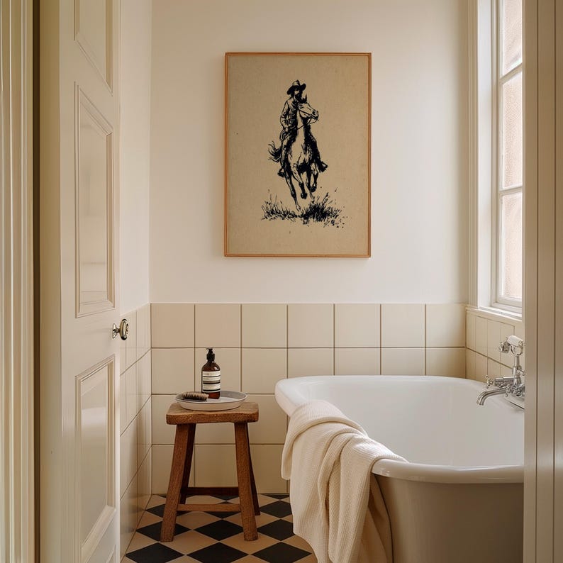 May include: A framed wall art print featuring a black ink illustration of a cowboy riding a horse on a tan background. The artwork is displayed on a wall above a white bathtub and a small wooden stool in a tiled bathroom setting.