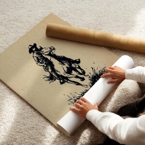 May include: A large square sheet of tan paper featuring a black ink illustration of a person riding a horse. The artwork depicts the rider and horse in motion, with textured lines and dark shading. A brown cardboard tube lies nearby on the surface.