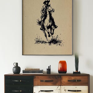 May include: A framed wall art print featuring a black ink illustration of a cowboy riding a galloping horse on a tan background. The artwork depicts the rider in a hat and the horse moving through tall grass, presented in a thin black frame.