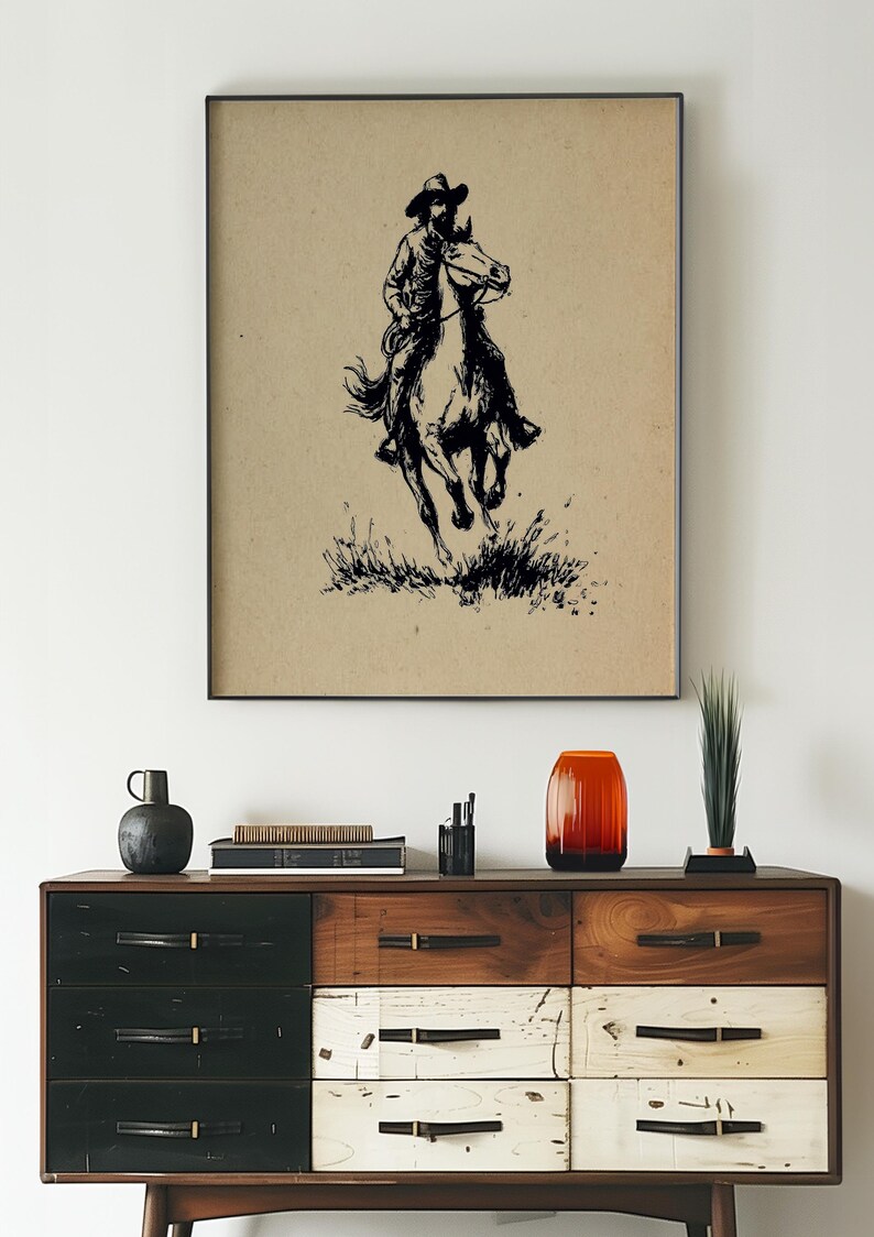May include: A framed wall art print featuring a black ink illustration of a cowboy riding a galloping horse on a tan background. The artwork depicts the rider in a hat and the horse moving through tall grass, presented in a thin black frame.