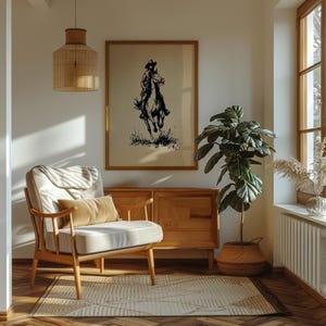 May include: A framed wall art print featuring a black ink illustration of a person riding a horse on a beige background. The artwork is displayed in a wooden frame, hanging above a wooden sideboard in a room with a light-colored armchair and a plant.